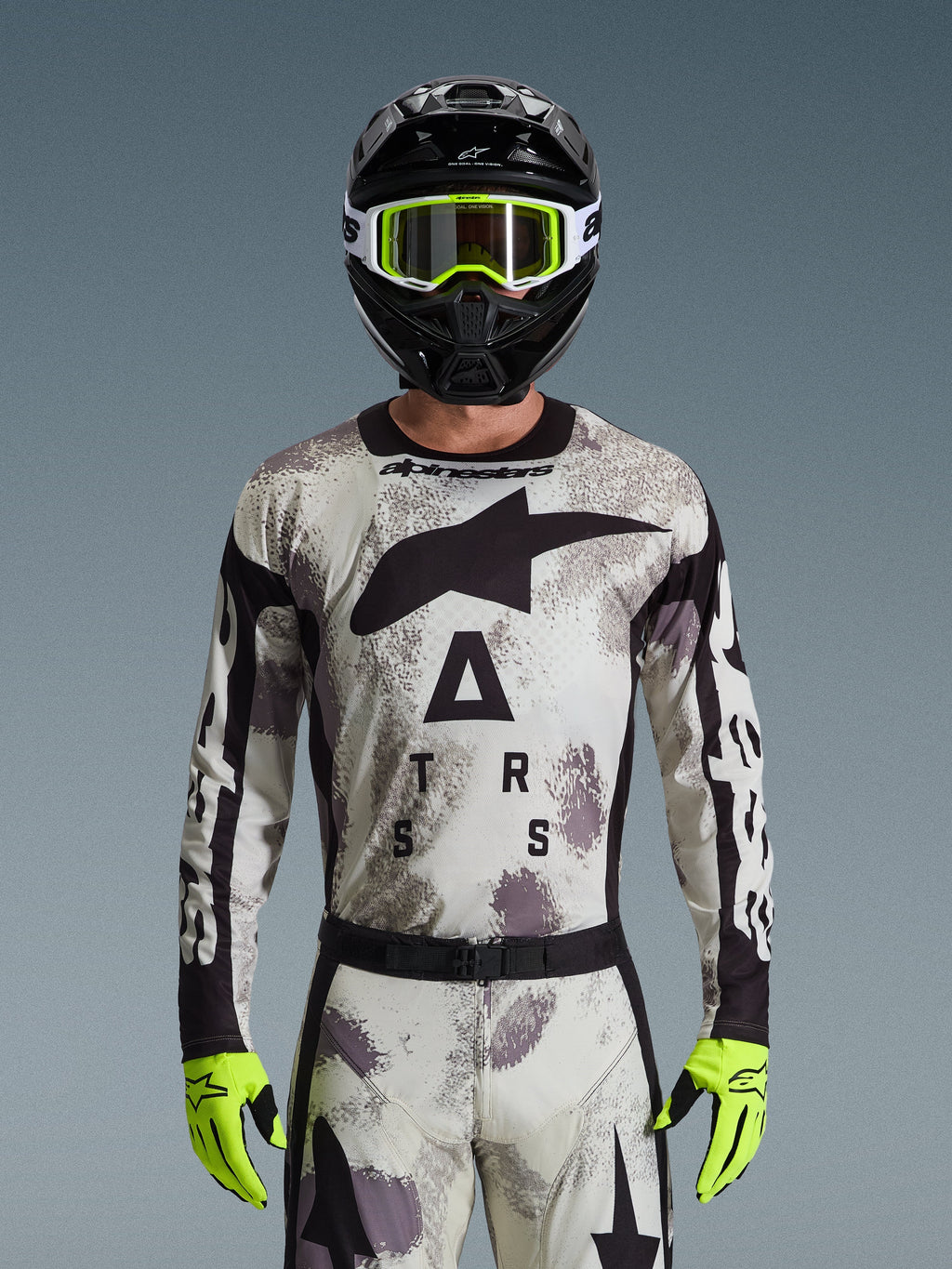 Alpinestars 2026 Racer Lahnd Jersey, conjunto de equipamento de motocross, padrão Sand Camo em cinzento e branco, logótipo de estrela preta grande e letras "ASTARS", usado com capacete preto, óculos amarelo néon e luvas amarelo néon, equipamento de corrida profissional de todo-o-terreno