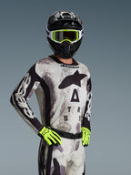 Piloto de motocross com camisola e conjunto de equipamento Alpinestars 2026 Racer Lahnd em Sand Camo, padrão de camuflagem cinzento claro com logótipo preto, capacete preto com óculos brancos e amarelo néon, luvas amarelo néon vibrante, vestuário profissional de corrida todo-o-terreno