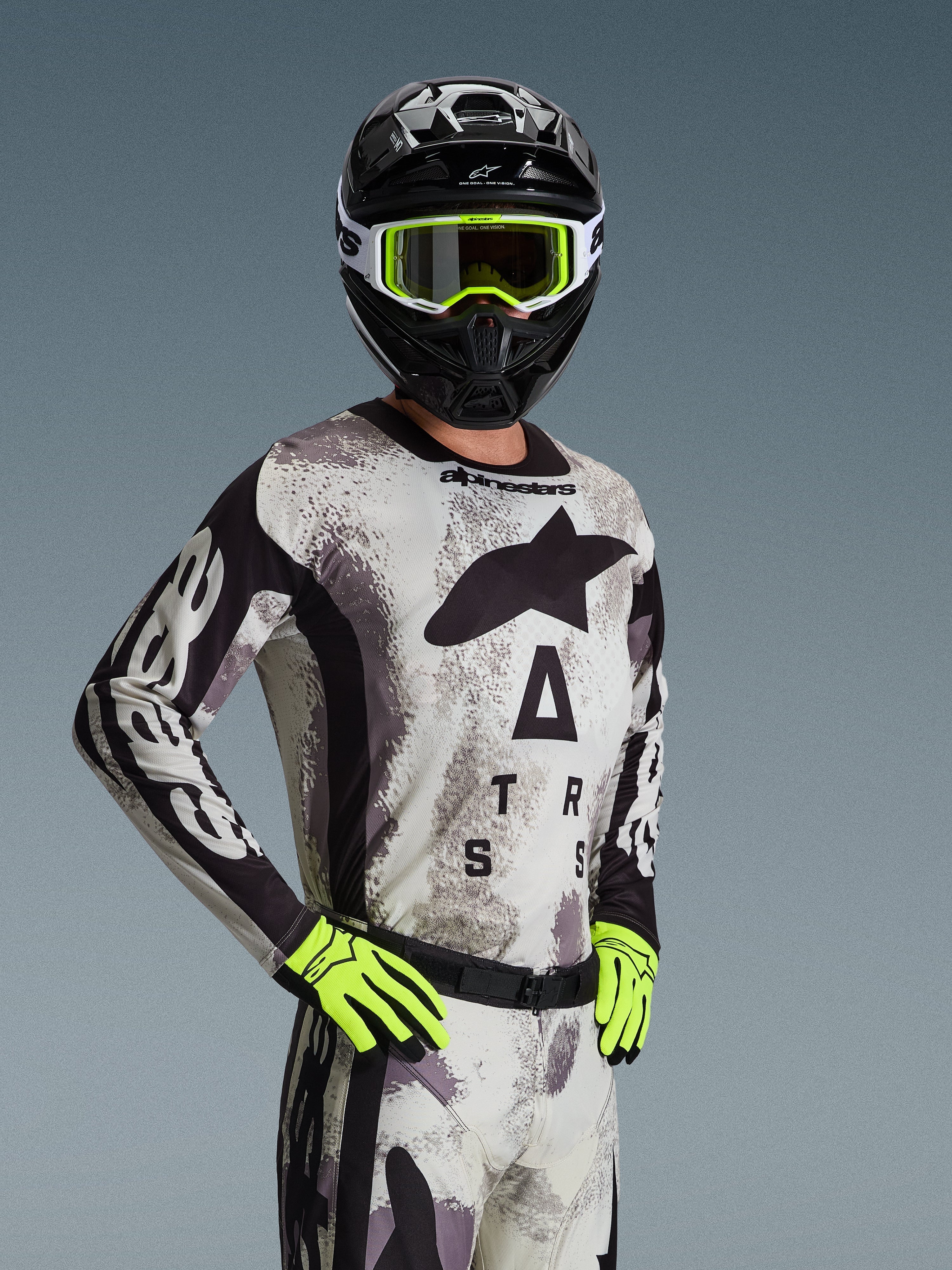 Piloto de motocross com camisola e conjunto de equipamento Alpinestars 2026 Racer Lahnd em Sand Camo, padrão de camuflagem cinzento claro com logótipo preto, capacete preto com óculos brancos e amarelo néon, luvas amarelo néon vibrante, vestuário profissional de corrida todo-o-terreno