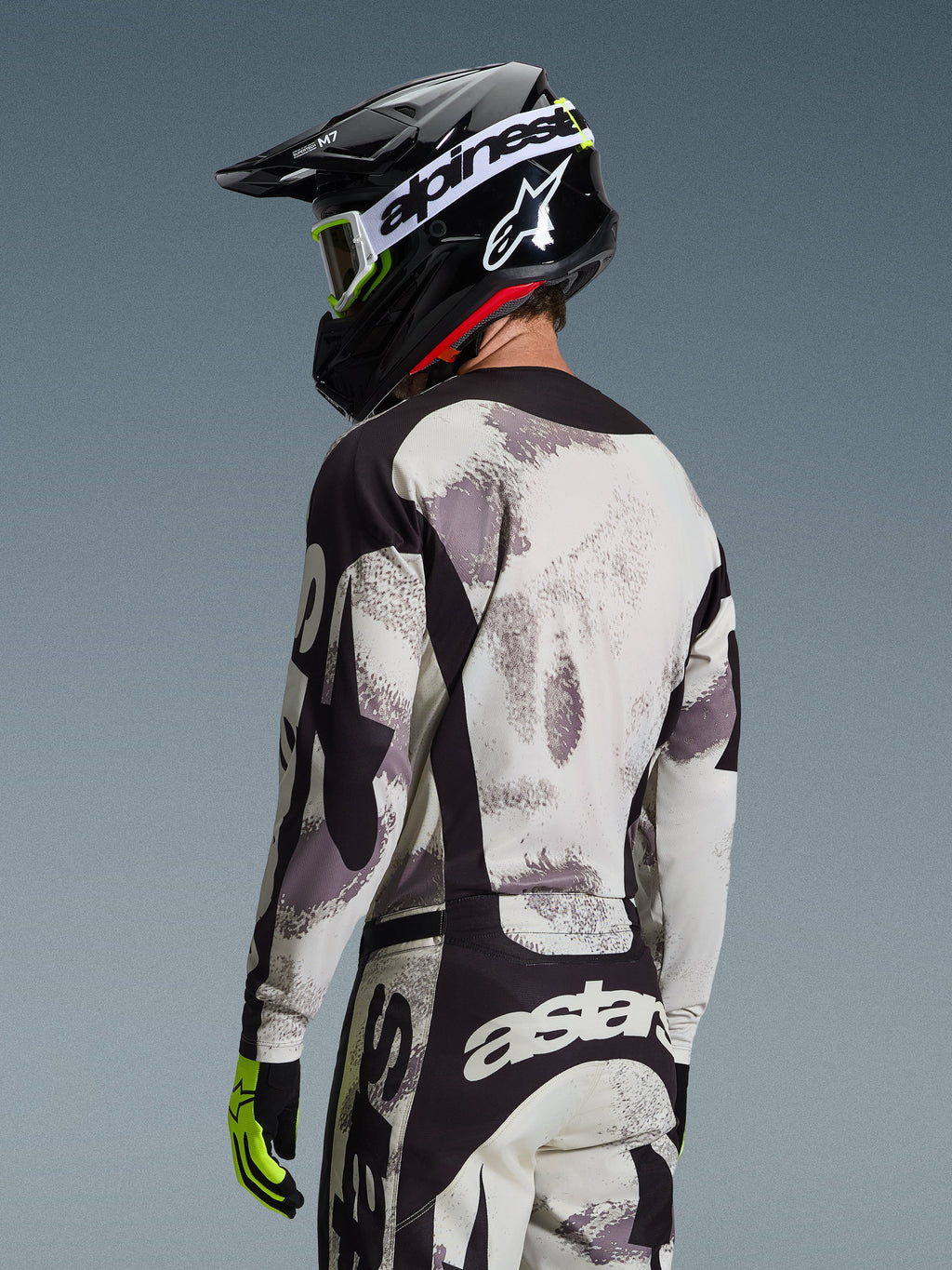 Piloto de motocross visto de costas a usar o Conjunto Racer Lahnd 2026 da Alpinestars, camisola e calças com padrão Sand Camo em cinzento e branco com detalhes pretos, capacete preto com logótipo Alpinestars branco, luvas amarelo néon e pretas, conjunto coordenado de performance para todo-o-terreno