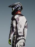 Piloto de motocross visto de costas a usar o Conjunto Racer Lahnd 2026 da Alpinestars, camisola e calças com padrão Sand Camo em cinzento e branco com detalhes pretos, capacete preto com logótipo Alpinestars branco, luvas amarelo néon e pretas, conjunto coordenado de performance para todo-o-terreno