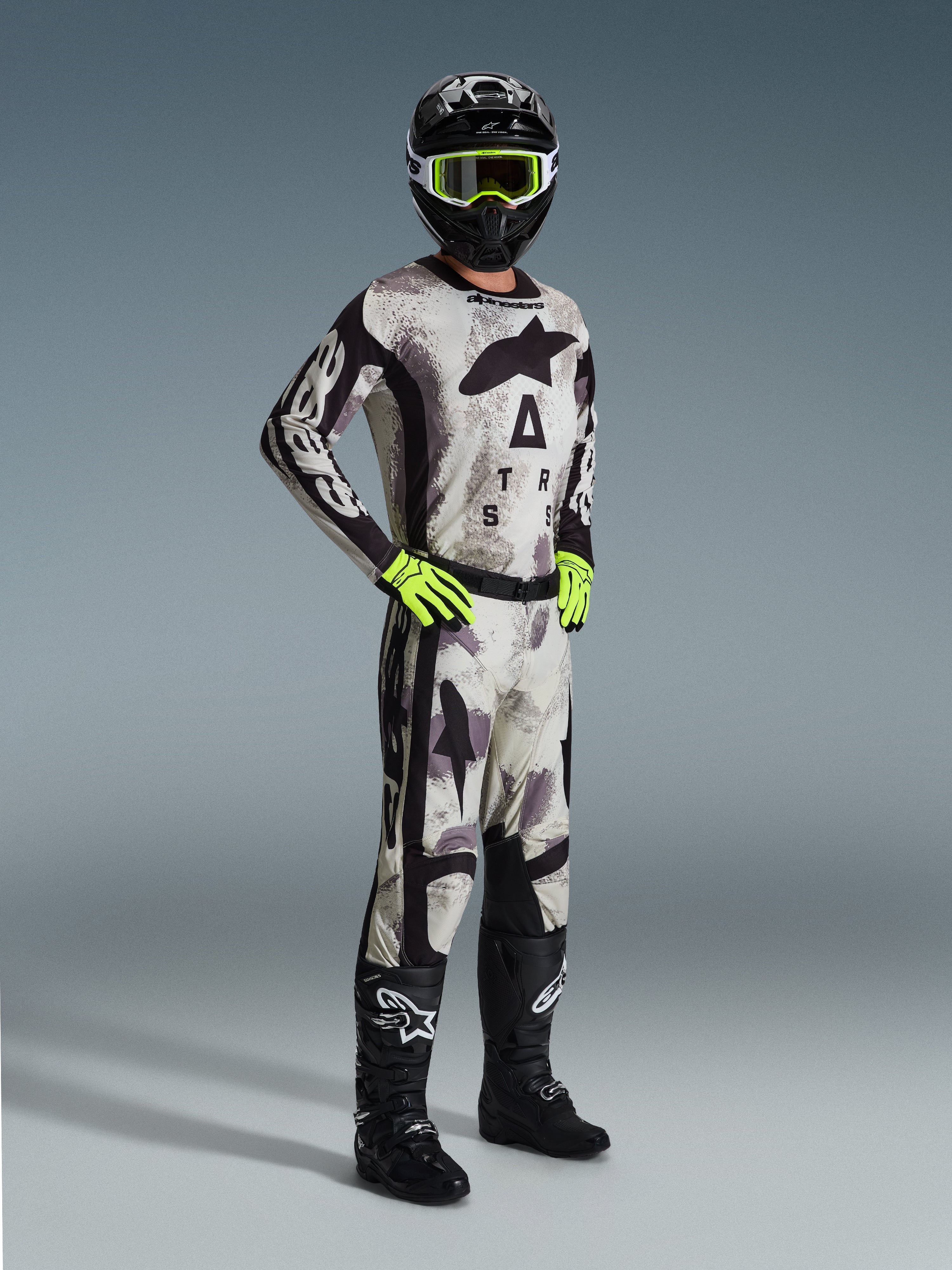 Alpinestars 2026 Racer Lahnd Gear Set, piloto de motocross com equipamento de corrida completo em Sand Camo e cinzento, camisola e calças a condizer com detalhes pretos e logótipos destacados, óculos e luvas em amarelo néon, capacete preto e botas pretas.