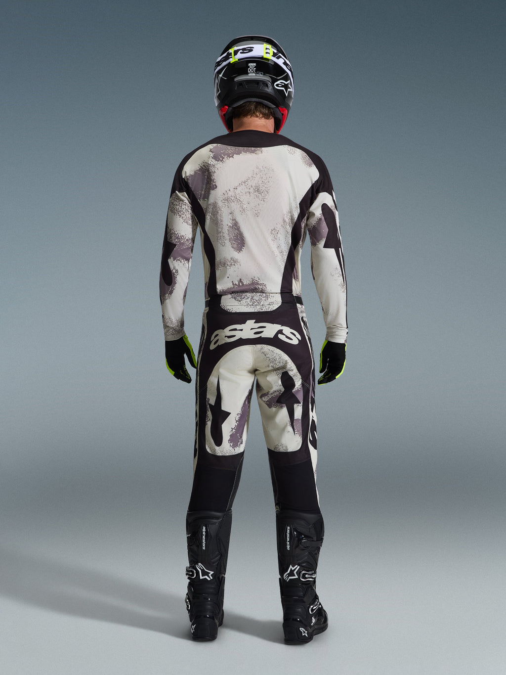 Piloto de motocross a usar o conjunto completo de equipamento Alpinestars 2026 Racer Lahnd, visto de costas, camisola e calças Sand Camo com padrão abstrato cinzento claro e preto, capacete preto, luvas pretas e amarelo néon, botas pretas, com a marca 'astars' na parte traseira das calças