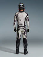 Piloto de motocross a usar o conjunto completo de equipamento Alpinestars 2026 Racer Lahnd, visto de costas, camisola e calças Sand Camo com padrão abstrato cinzento claro e preto, capacete preto, luvas pretas e amarelo néon, botas pretas, com a marca 'astars' na parte traseira das calças