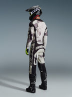 Piloto de motocross a usar as calças Alpinestars 2026 Racer Lahnd em Sand Camo e cinzento, conjunto completo de equipamento com camisola a condizer, capacete preto e botas pretas, vista traseira realçando o branding arrojado e o design ergonómico.