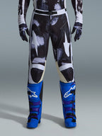 Alpinestars 2026 Racer Lahnd Pants, calças de motocross, Iron Camo, usadas com luvas camufladas a combinar e botas azul vibrante e roxas, com grafismos de estrelas brancas estilizadas num padrão camuflado escuro, fecho de fivela magnética técnica, equipamento profissional de corrida para todo-o-terreno