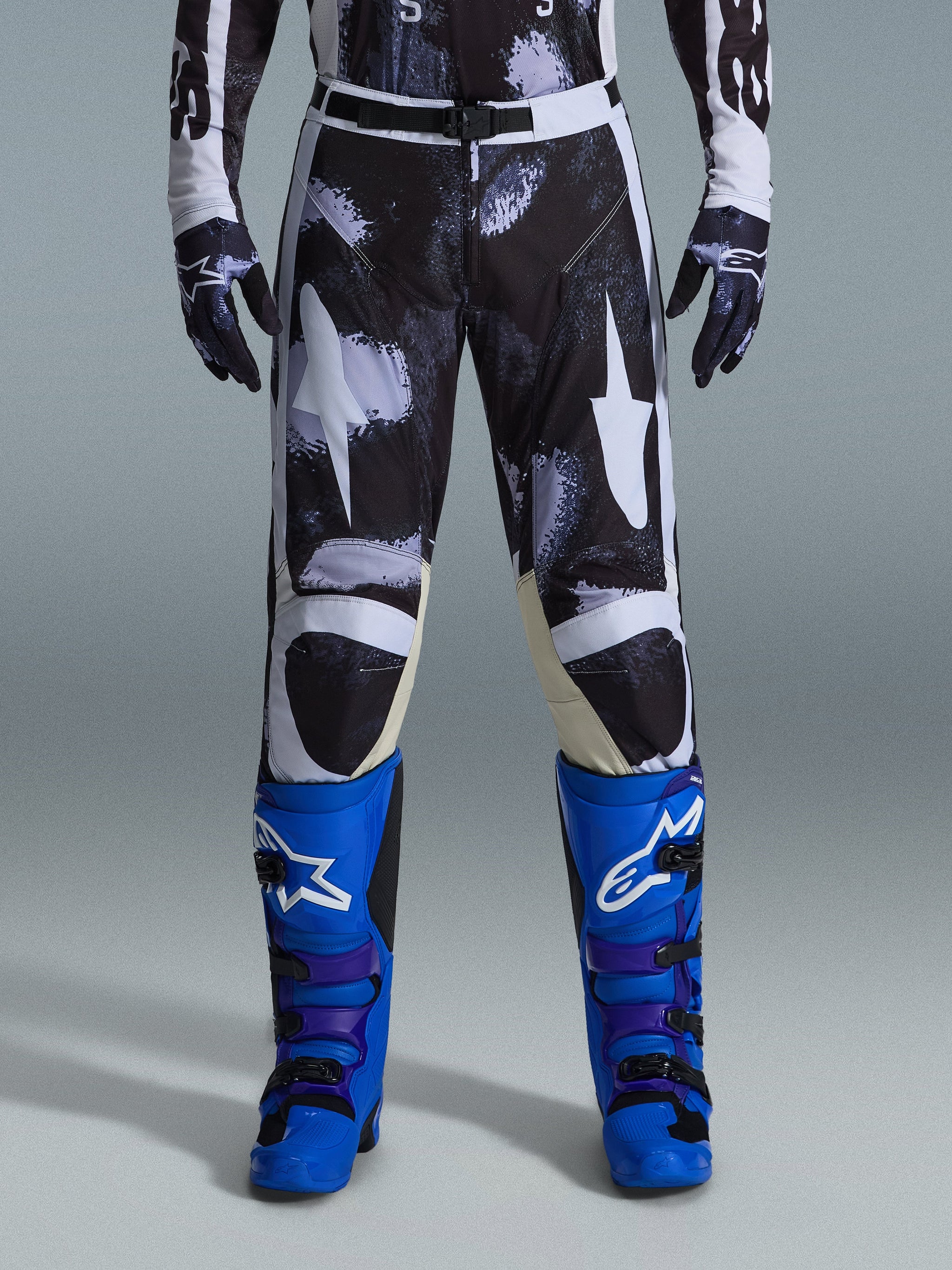 Alpinestars 2026 Racer Lahnd Pants, calças de motocross, Iron Camo, usadas com luvas camufladas a combinar e botas azul vibrante e roxas, com grafismos de estrelas brancas estilizadas num padrão camuflado escuro, fecho de fivela magnética técnica, equipamento profissional de corrida para todo-o-terreno