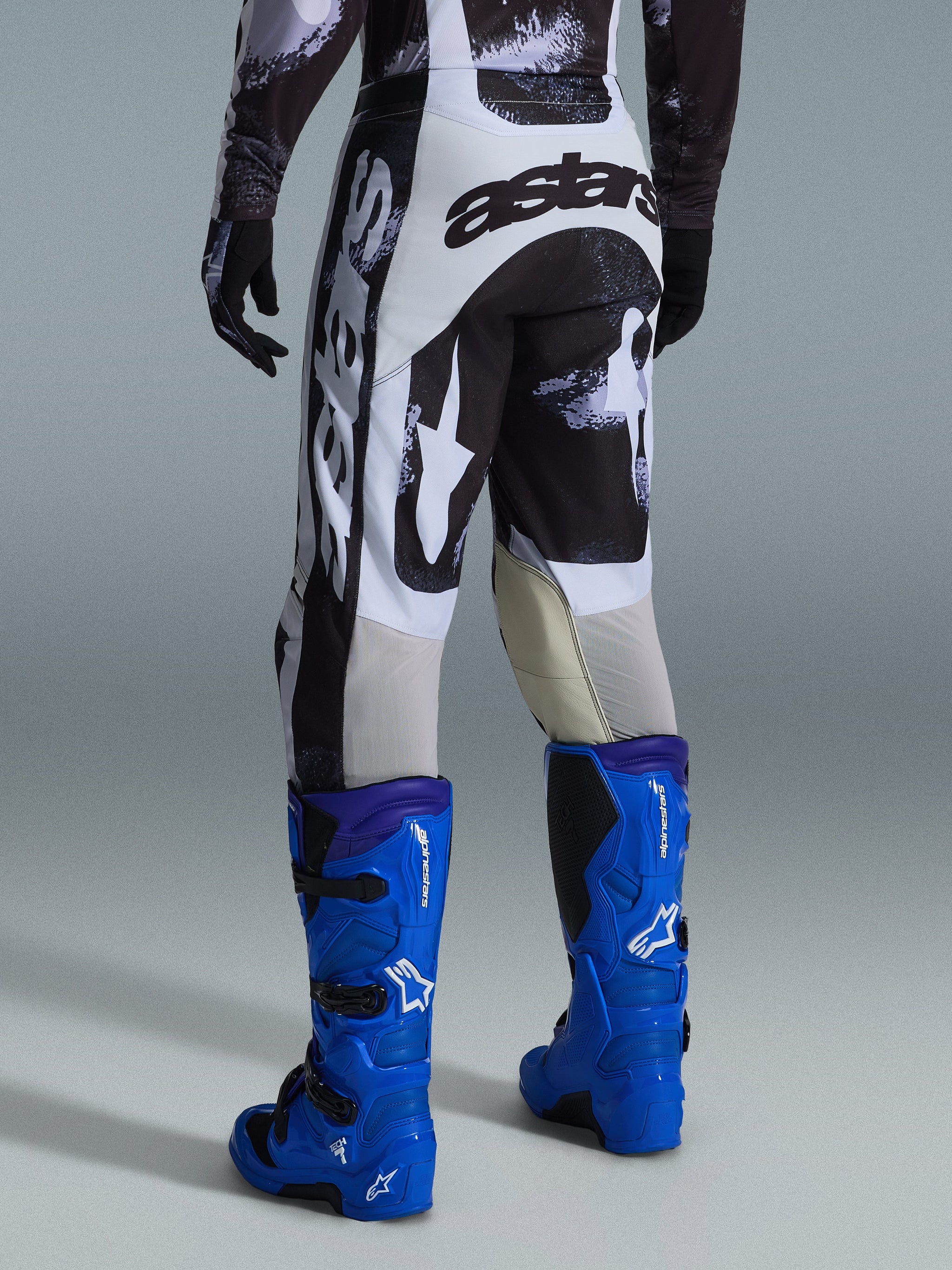 Alpinestars 2026 Racer Lahnd Pants, calças de motocross, padrão camuflado Iron Camo em cinzento e preto, vista traseira com grande logótipo astars a branco e preto, combinadas com botas de motocross em azul brilhante, equipamento profissional de corrida para todo-o-terreno