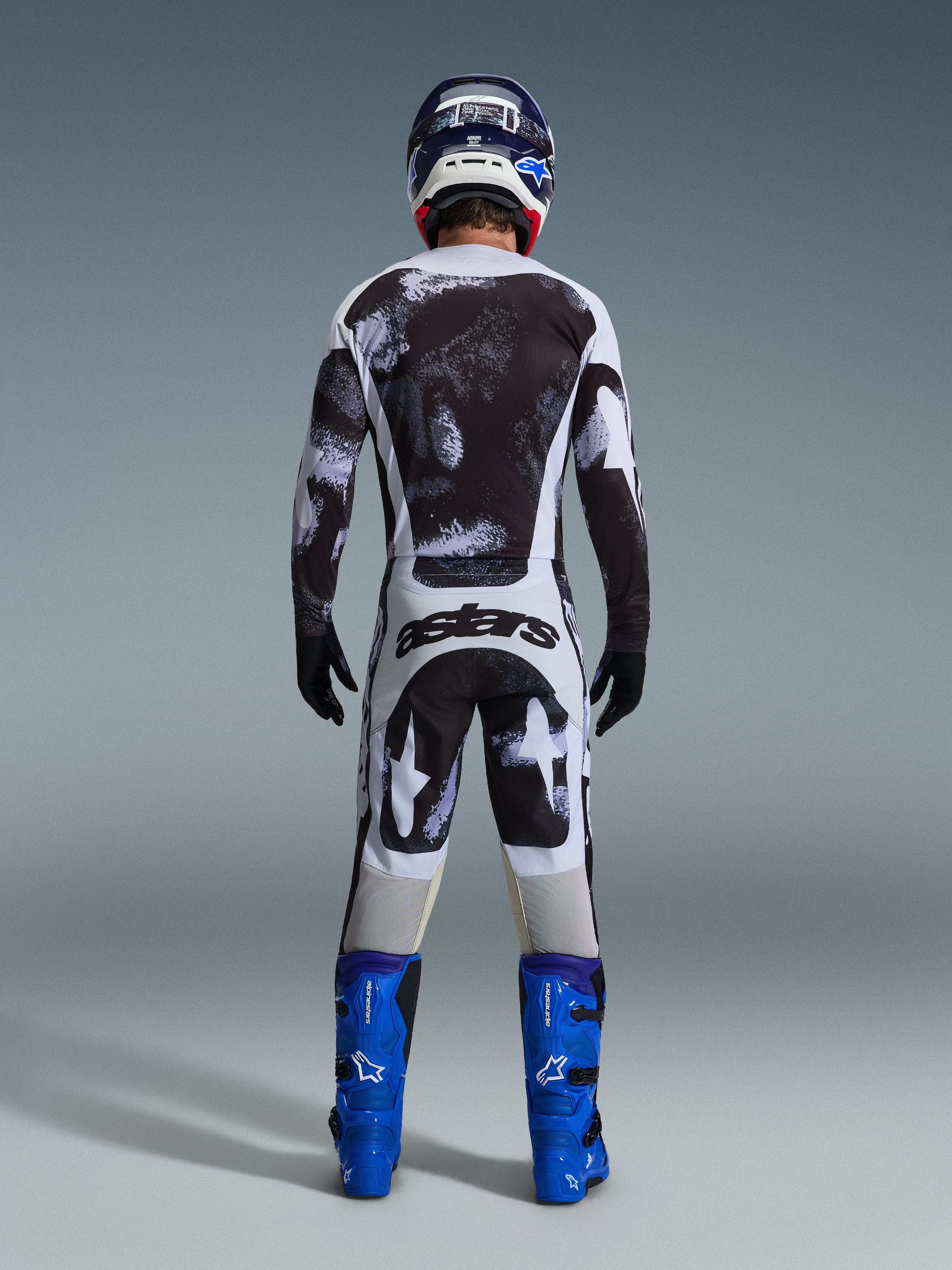 Piloto de motocross visto de costas a usar a camisola Alpinestars 2026 Racer Lahnd e calças a condizer em Iron Camo, com um padrão de salpicos em preto e branco, botas azul vivo e capacete coordenado, conjunto completo de equipamento para corridas off-road