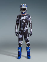Piloto de motocross a usar calças Alpinestars 2026 Racer Lahnd em Iron Camo, com padrão de camuflagem cinzento e preto, camisola a condizer, capacete azul e branco com óculos, e botas azul vibrante, conjunto de equipamento de motocross coordenado.