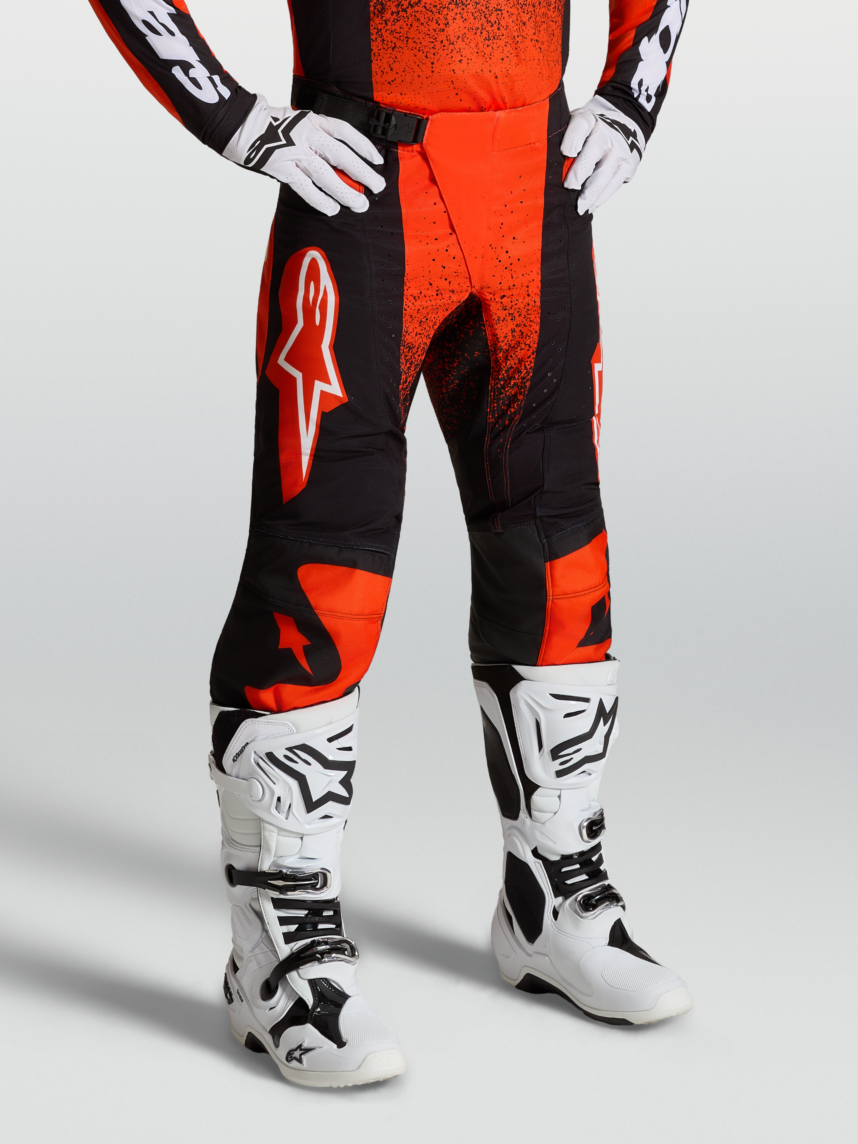 Alpinestars Calças Techstar Swan, calças de motocross para homem, vermelhas e pretas, com painéis elásticos e grafismos brancos, vestidas com botas de moto brancas.