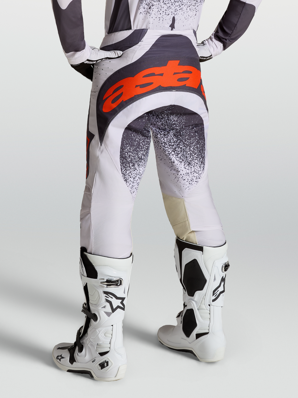 Alpinestars Calças Techstar Swan, calças de motocross para homem, cinzento e lilás com detalhes em vermelho fluo, vista traseira com botas Tech 10