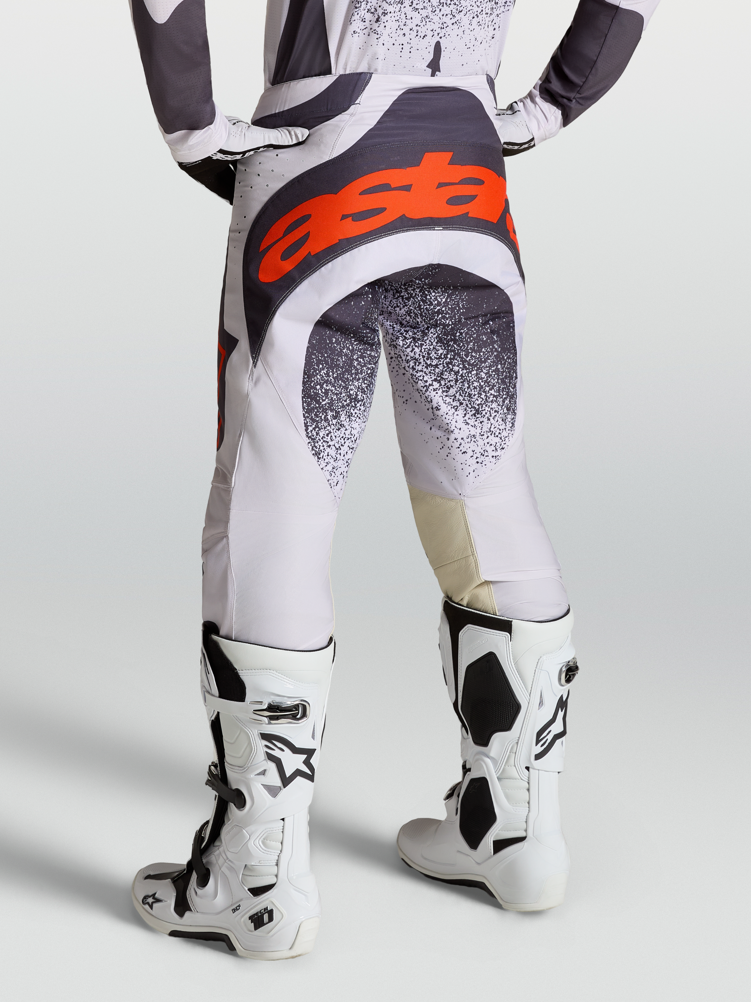Alpinestars Calças Techstar Swan, calças de motocross para homem, cinzento e lilás com detalhes em vermelho fluo, vista traseira com botas Tech 10