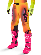 Alpinestars 2025 Supertech Lipan Gear Set, calças de motocross, gradiente em amarelo e cor de laranja com pormenores em cor-de-rosa e roxo, com logótipos Alpinestars pretos destacados nas coxas, usadas com luvas amarelas e botas de motocross cor-de-rosa vibrante