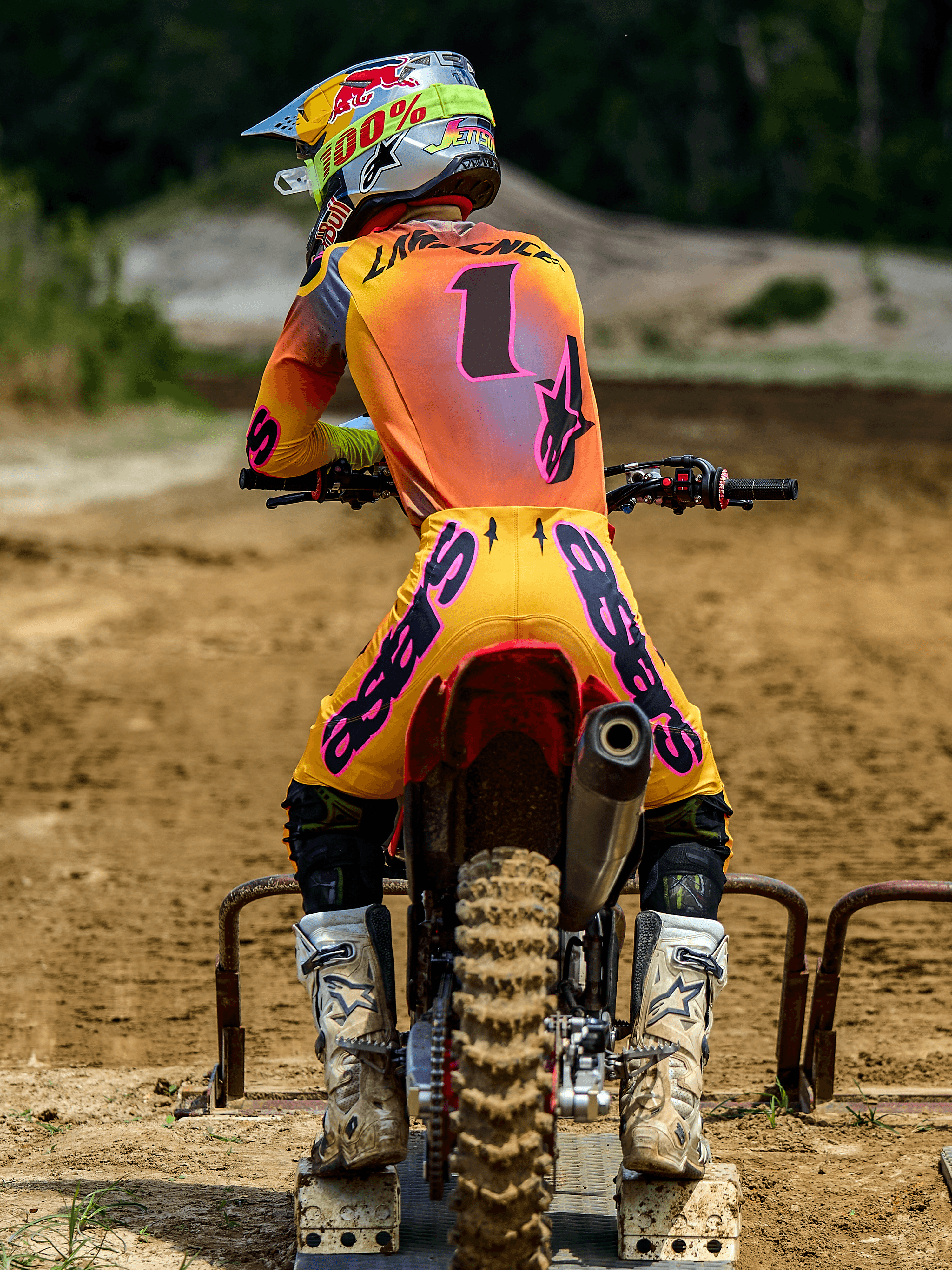 Piloto de motocross visto de costas a usar o Conjunto de Equipamento Alpinestars Supertech Lipan 2025, amarelo e rosa, camisola com o número 1 e 'LAWRENCE' nas costas, calças amarelas com a marca 'ASTARS' em rosa vibrante, botas brancas e capacete a condizer, sentado numa mota de cross no portão de partida.