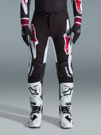 Alpinestars 2026 Supertech AFD Vista Pants, calças de motocross, pretas, vermelhas e brancas, com grandes logótipos gráficos nas coxas e joelhos, usadas com botas de competição brancas e luvas pretas, design desportivo de corrida para competição off-road de elite