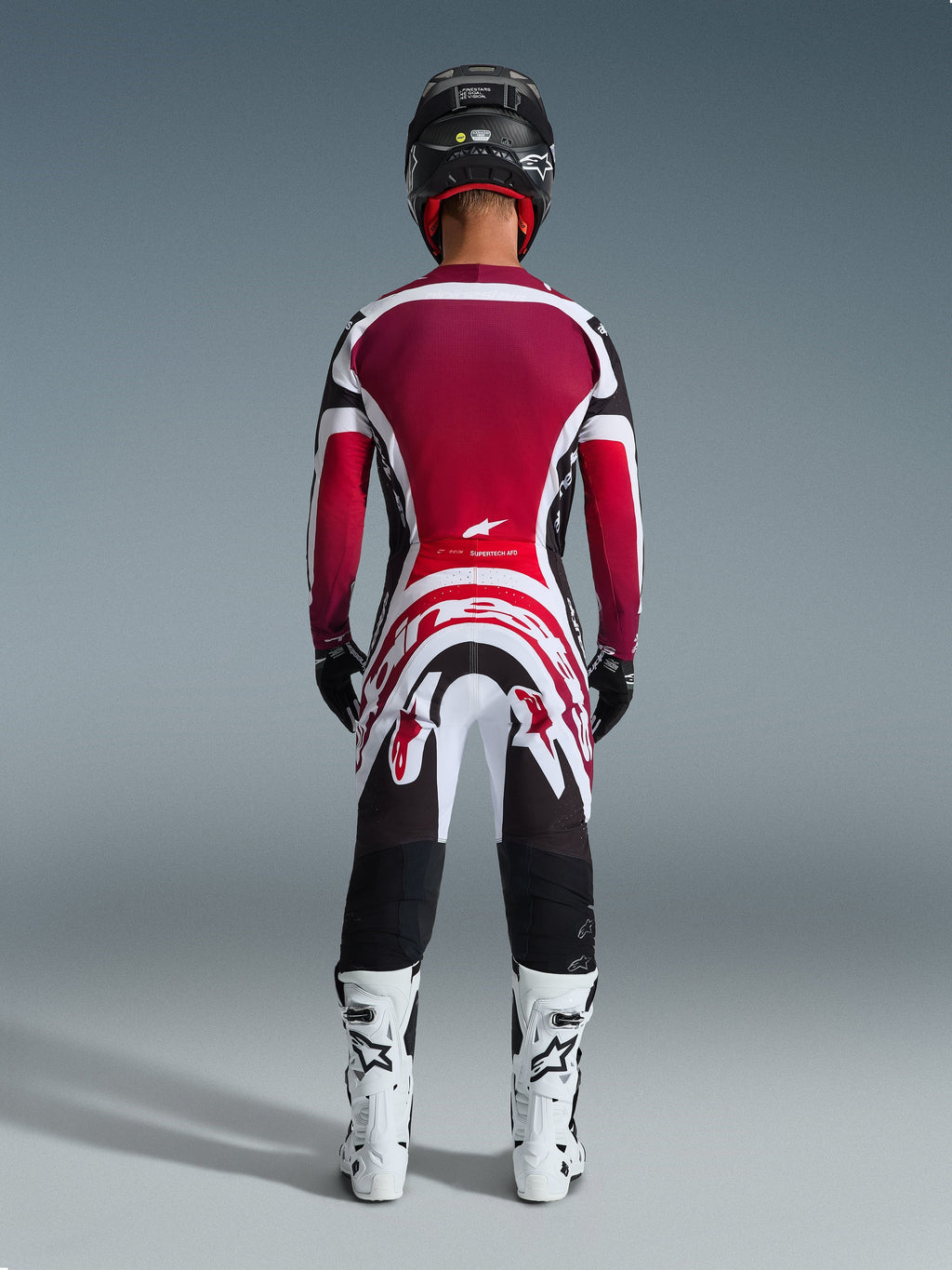 Piloto de motocross visto de trás a usar calças Alpinestars 2026 Supertech AFD Vista, Pretas, Vermelhas e Brancas, combinadas com uma camisola a condizer, botas de competição brancas e capacete preto, conjunto de equipamento profissional completo para corridas off-road