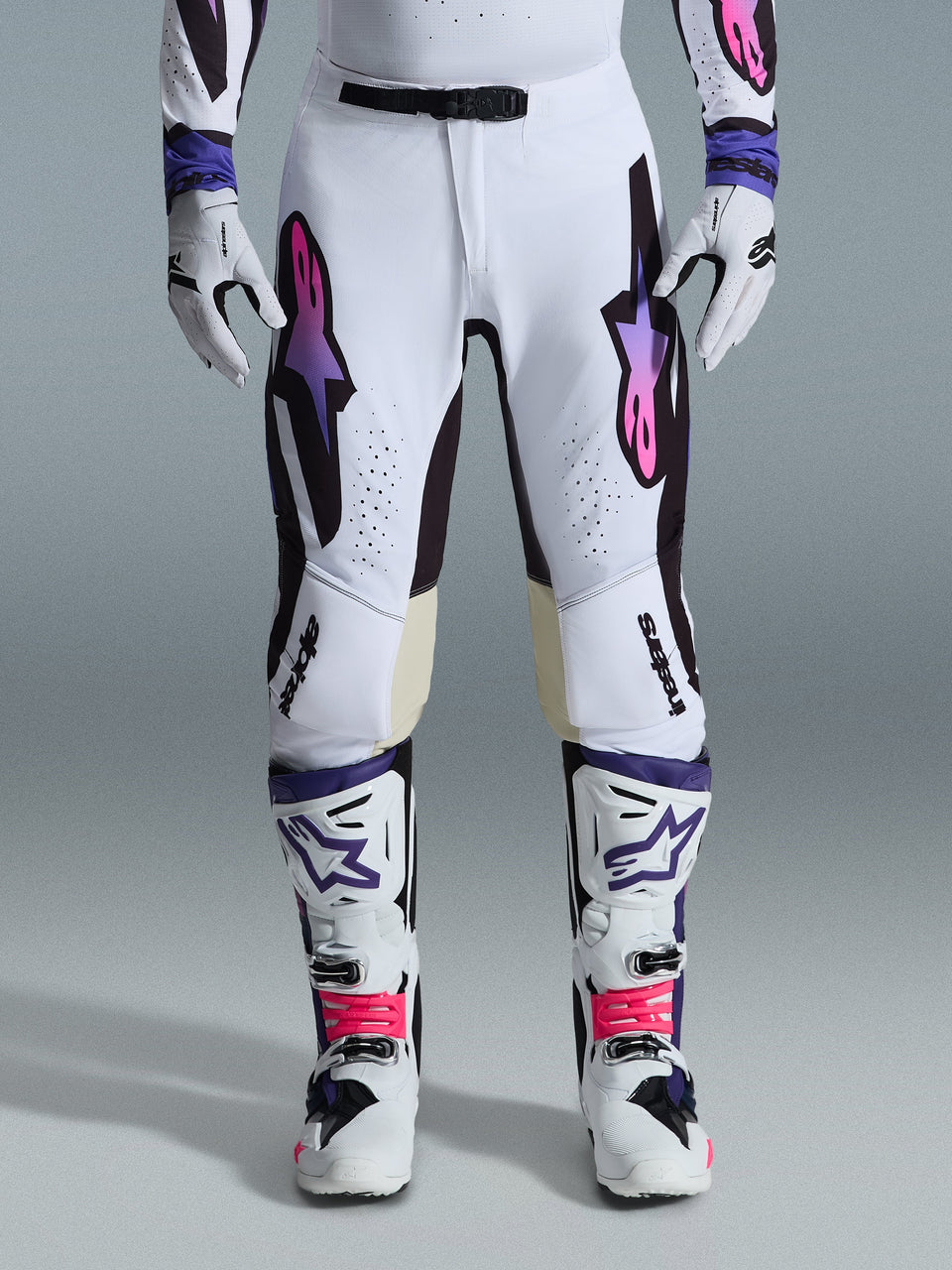 Alpinestars 2026 Supertech AFD Vista Pants, calças de motocross, brancas, roxas e pretas, usadas com botas e luvas brancas a condizer, com logótipos Astars em degradê de rosa para roxo nas coxas, design de competição profissional para performance off-road.
