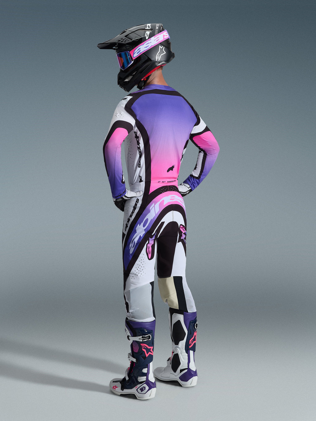 Piloto de motocross visto de costas a usar as calças Alpinestars 2026 Supertech AFD Vista, na cor Branco, Roxo e Preto, combinadas com uma camisola em gradiente roxo e rosa a condizer, capacete preto e botas de competição brancas e roxas para um visual completo de corrida off-road.