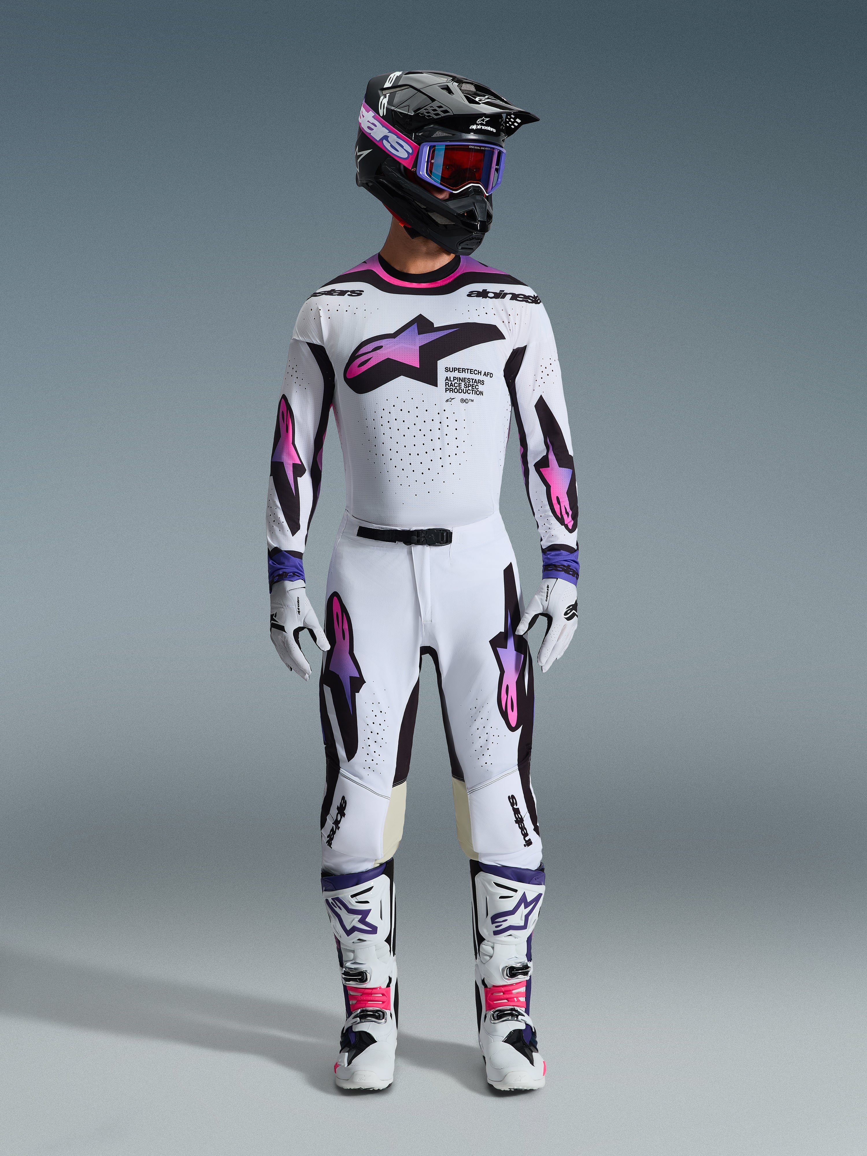 Piloto de motocross com um conjunto completo de equipamento de corrida, calças e camisola Alpinestars Supertech AFD Vista 2026 em branco, roxo e preto, luvas a condizer, botas brancas e pretas, capacete preto com óculos roxos, ajuste atlético para performance em todo-o-terreno