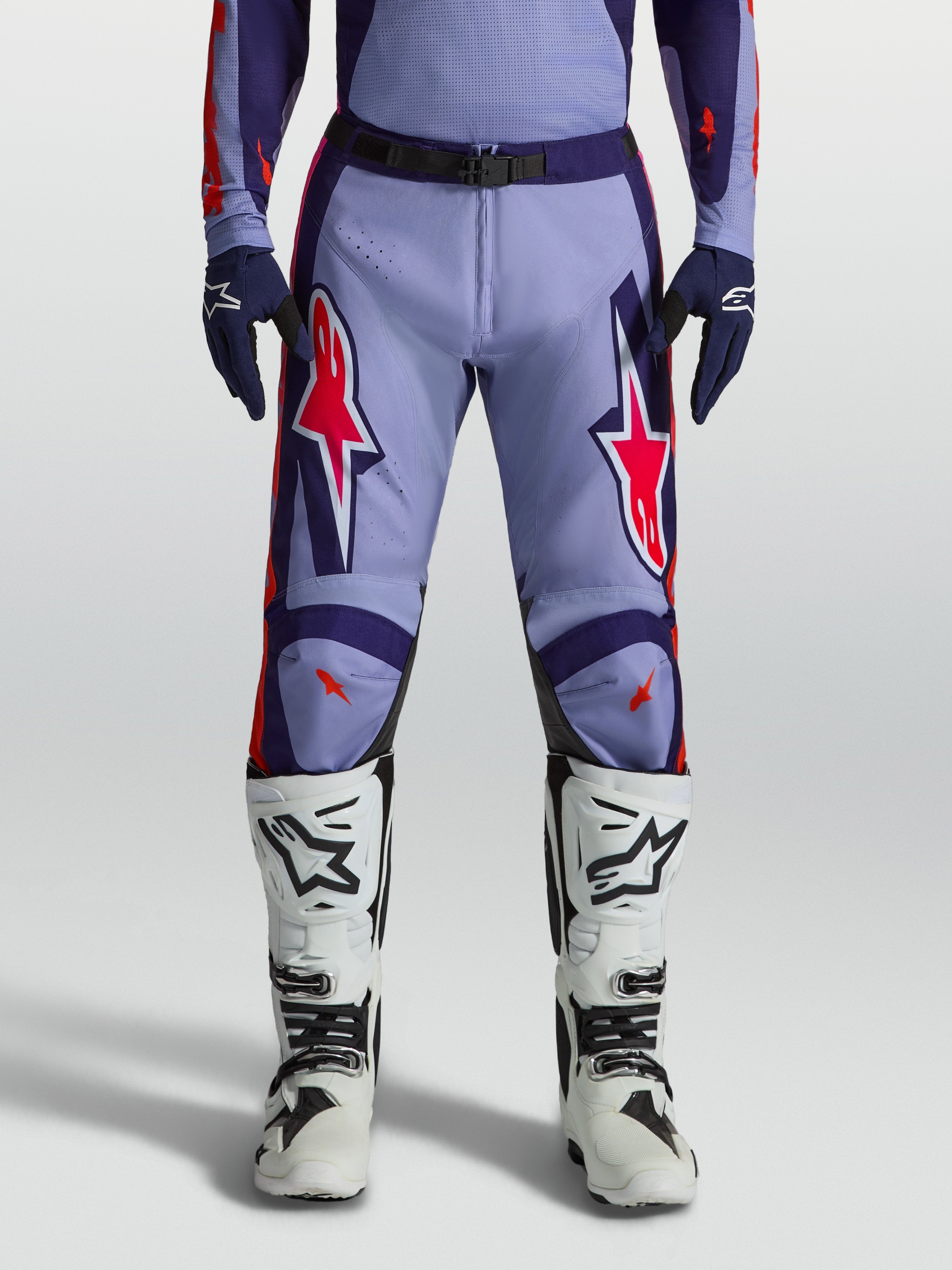Alpinestars calças Racer Air Hunt, calças de motocross em violeta claro e roxo profundo com detalhes rosa fluorescente, design ventilado, usadas com botas brancas.