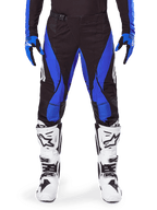 Alpinestars 2025 Techstar Dreem Pants, calças de motocross, pretas e azuis, usadas com camisola e luvas a condizer e botas brancas, com painéis laterais azuis e corte atlético de corrida, material durável para corridas off-road