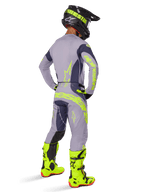 Piloto de motocross visto de costas a usar equipamento completo de corrida, incluindo calças Alpinestars 2025 Techstar Dreem em cinzento e cinzento escuro com detalhes vibrantes em amarelo néon, camisola cinzenta a condizer com grafismos néon, botas amarelo néon brilhante e capacete preto, conjunto de motocross profissional coordenado