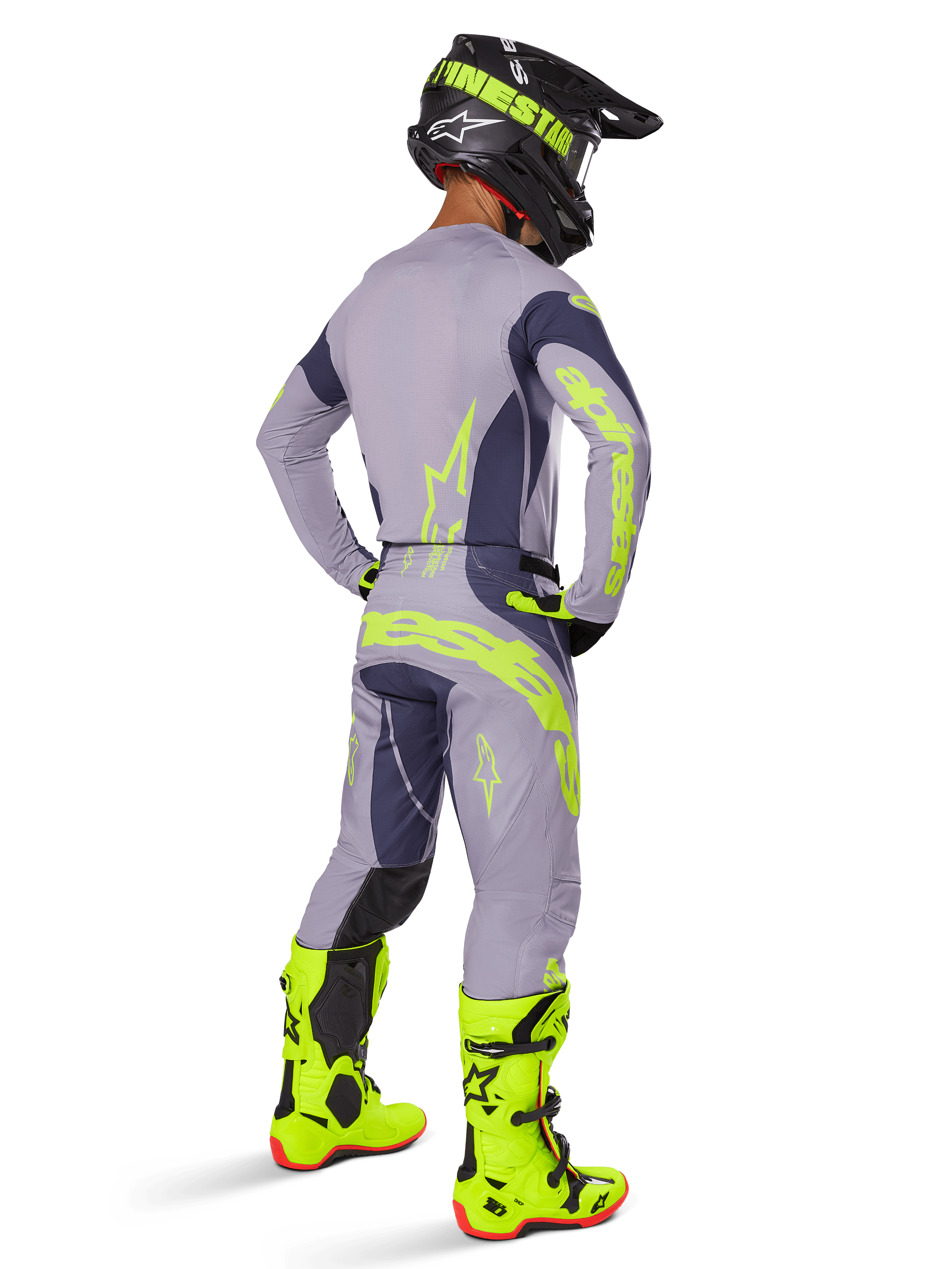 Piloto de motocross visto de costas a usar equipamento completo de corrida, incluindo calças Alpinestars 2025 Techstar Dreem em cinzento e cinzento escuro com detalhes vibrantes em amarelo néon, camisola cinzenta a condizer com grafismos néon, botas amarelo néon brilhante e capacete preto, conjunto de motocross profissional coordenado