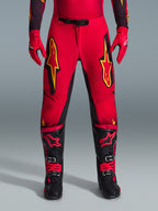Alpinestars 2026 Supertech Scenz Pants, calças de motocross, vermelhas, pretas e amarelas, ajuste atlético de nível profissional com grandes logótipos de estrela amarelos nas coxas, combinadas com luvas vermelhas a condizer e botas técnicas pretas e vermelhas, concebidas para performance de elite em corridas todo-o-terreno