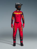 Calças Alpinestars 2026 Supertech Scenz, calças de motocross, vermelhas, pretas e amarelas, vestidas por um piloto num conjunto completo de competição, camisola vermelha e preta com logótipos amarelos, capacete preto, luvas vermelhas e botas pretas, design atlético pro-fit para competição todo-o-terreno