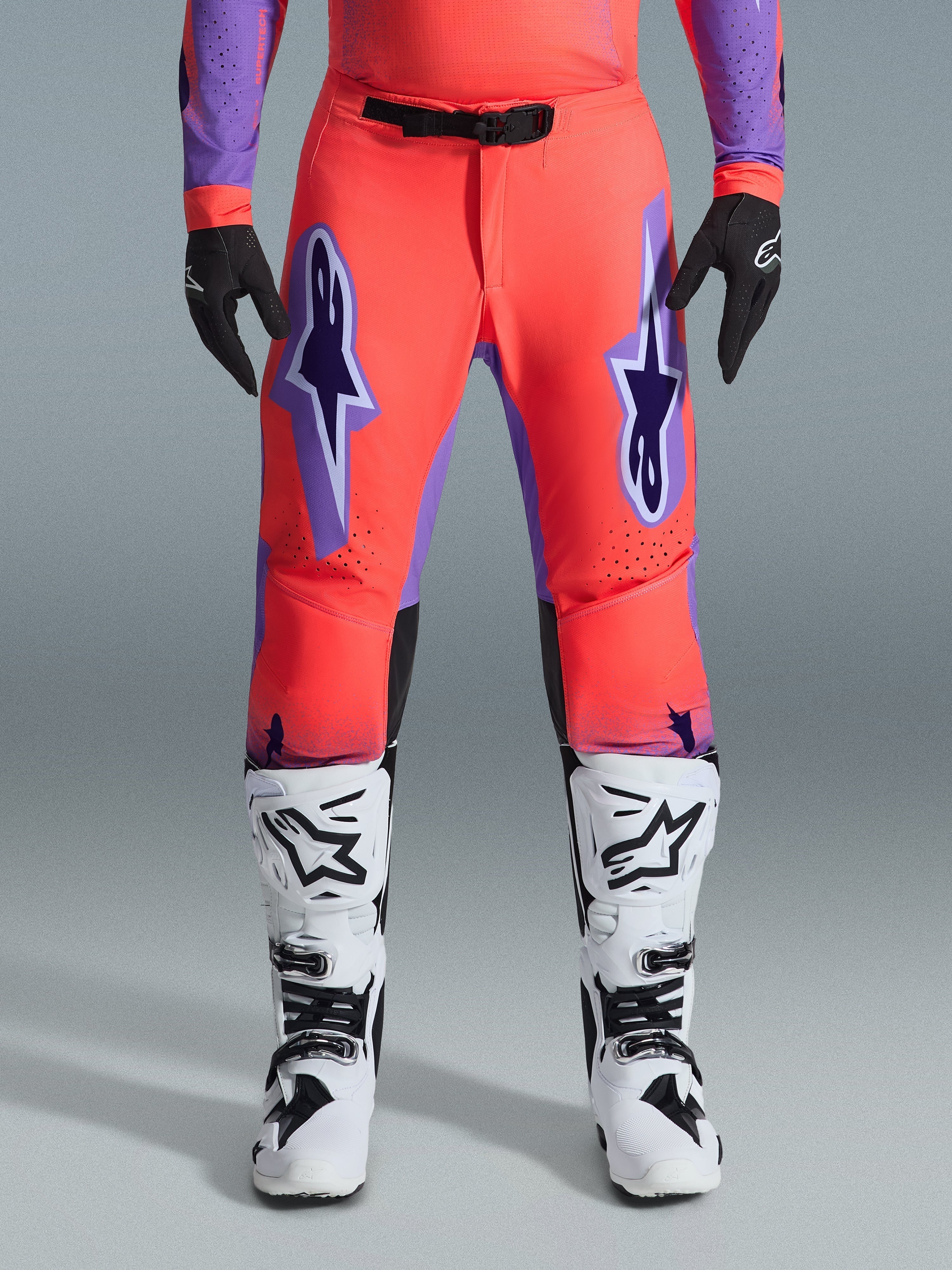 Alpinestars 2026 Supertech Scenz Pants, calças de motocross, coral vibrante e roxo, usadas com camisola a condizer, botas brancas e pretas e luvas pretas, com grandes logótipos Astars roxos nas coxas e fivela magnética preta na cintura, ajuste atlético profissional para corrida