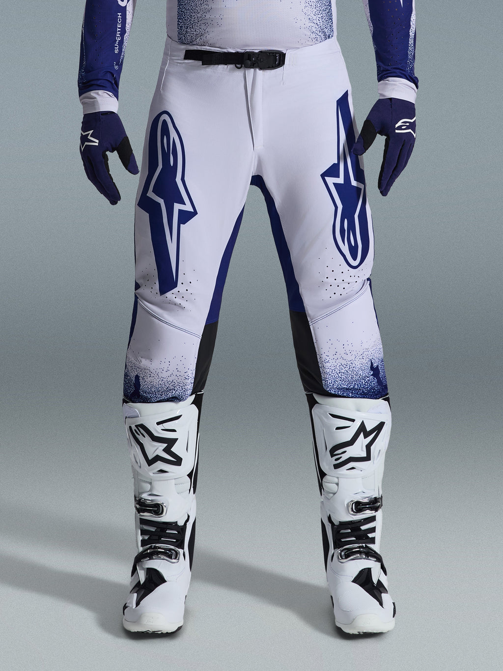 Alpinestars 2026 Supertech Scenz Pants, calças de motocross, cinzento claro e azul escuro, usadas com botas de motocross brancas e pretas e luvas azul escuro, com grandes logótipos Alpinestars azuis nas coxas, fivela magnética SX1 e ajuste atlético para corridas de todo-o-terreno