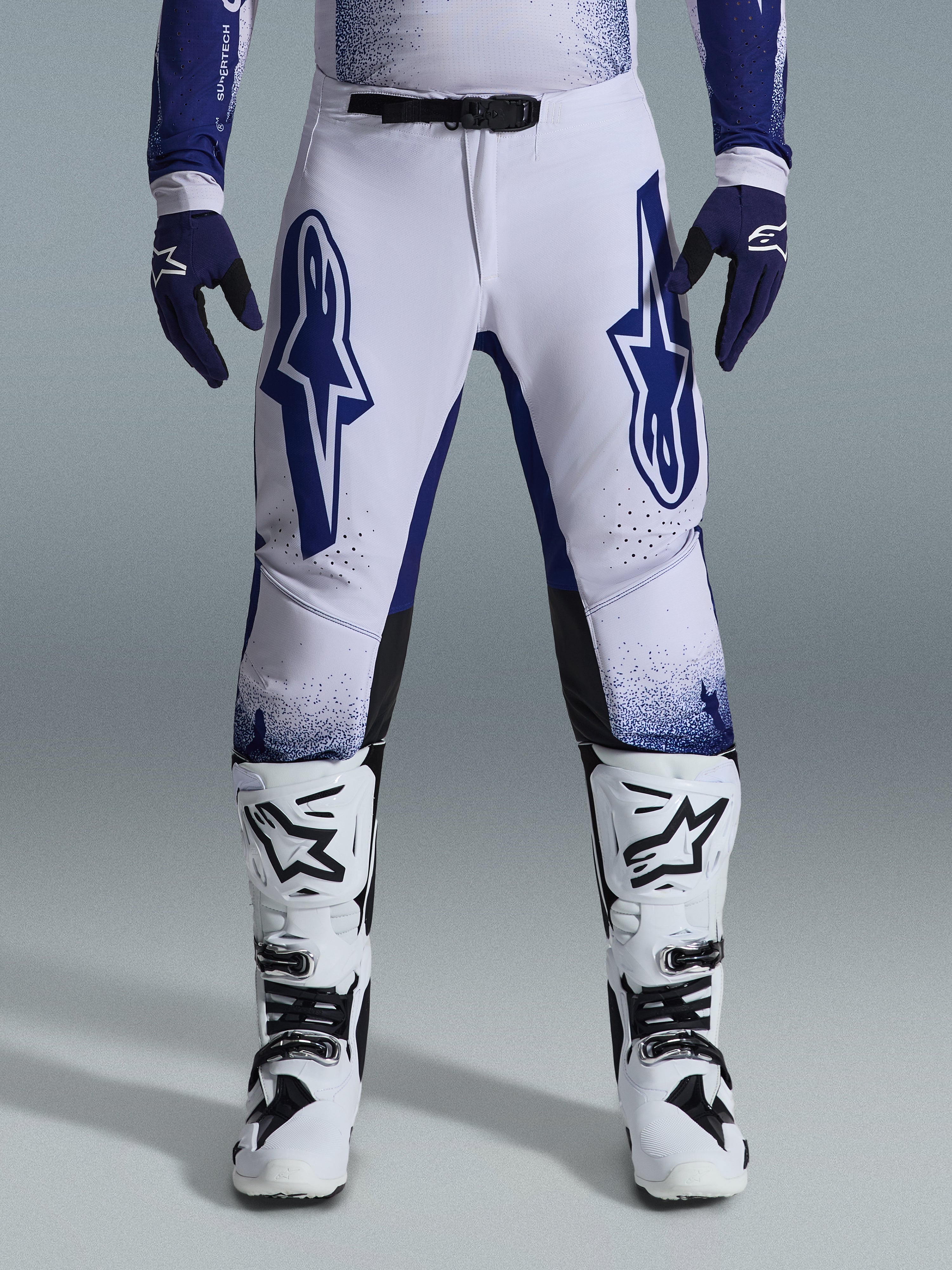 Alpinestars 2026 Supertech Scenz Pants, calças de motocross, cinzento claro e azul escuro, usadas com botas de motocross brancas e pretas e luvas azul escuro, com grandes logótipos Alpinestars azuis nas coxas, fivela magnética SX1 e ajuste atlético para corridas de todo-o-terreno