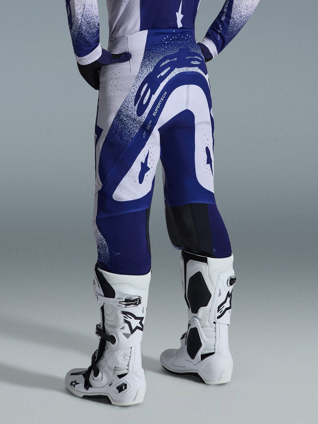 Alpinestars 2026 Supertech Scenz Pants, calças de motocross, cinzento claro e azul escuro, vista traseira, usadas com camisola a condizer e botas brancas, com grafismos azuis marcantes e detalhes salpicados, ajuste atlético de alta performance para corridas todo-o-terreno