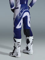 Alpinestars 2026 Supertech Scenz Pants, calças de motocross, cinzento claro e azul escuro, vista traseira, usadas com camisola a condizer e botas brancas, com grafismos azuis marcantes e detalhes salpicados, ajuste atlético de alta performance para corridas todo-o-terreno