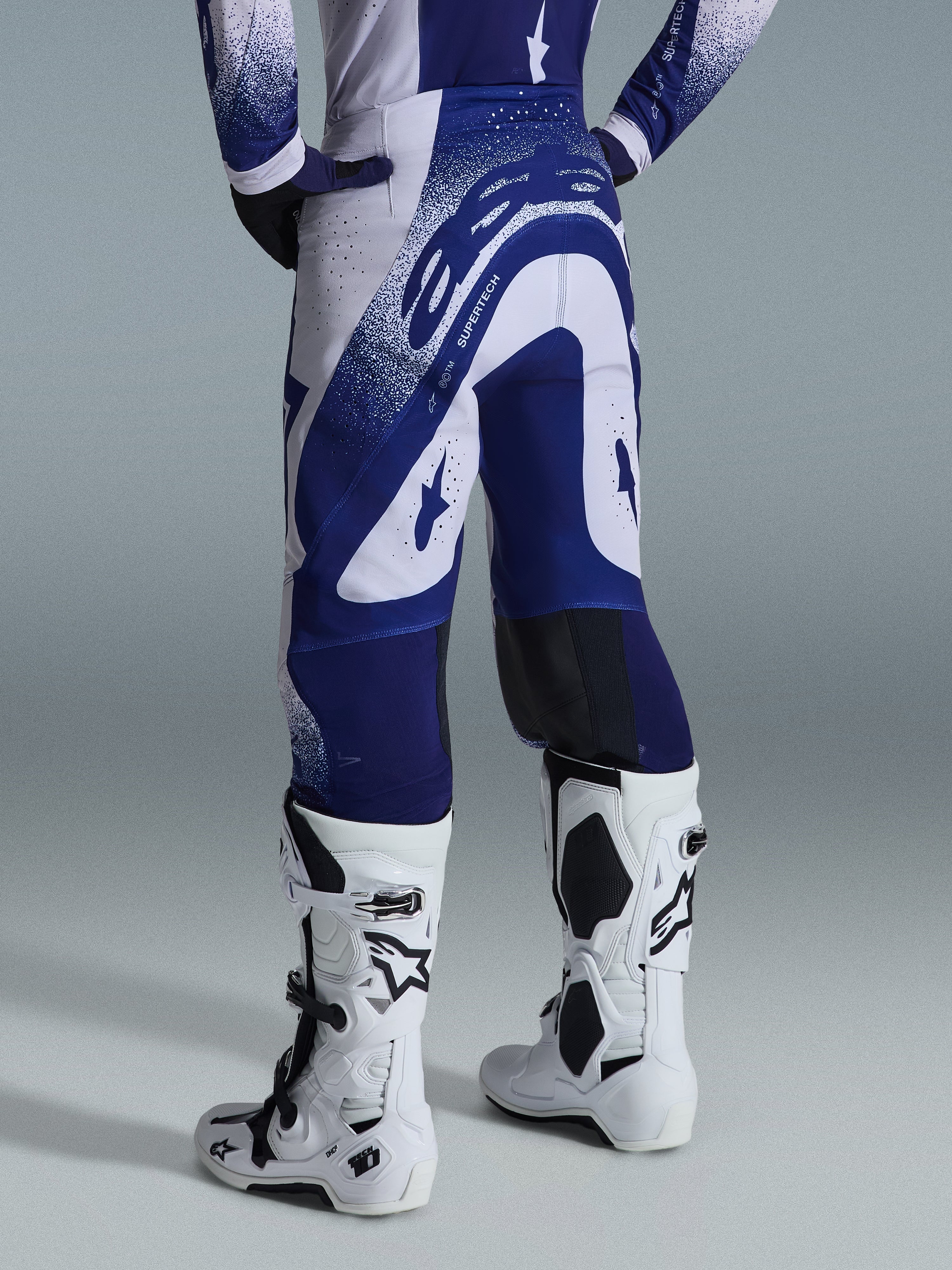 Alpinestars 2026 Supertech Scenz Pants, calças de motocross, cinzento claro e azul escuro, vista traseira, usadas com camisola a condizer e botas brancas, com grafismos azuis marcantes e detalhes salpicados, ajuste atlético de alta performance para corridas todo-o-terreno