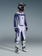 Piloto de motocross vestido com as Calças Supertech Scenz 2026 em cinzento claro e azul escuro, camisola azul e branca a condizer com a marca 'astars' e efeito de pintura em spray, capacete preto com óculos de proteção brancos, botas de motocross brancas e pretas, equipamento profissional de corrida todo-o-terreno coordenado.