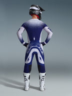Alpinestars 2026 Supertech Scenz Pants, calças de motocross, cinzento claro e azul escuro, vista traseira de um piloto a usar um conjunto de equipamento a condizer com gráficos de gradiente salpicado, botas brancas e capacete preto, ajuste atlético profissional para corridas todo-o-terreno