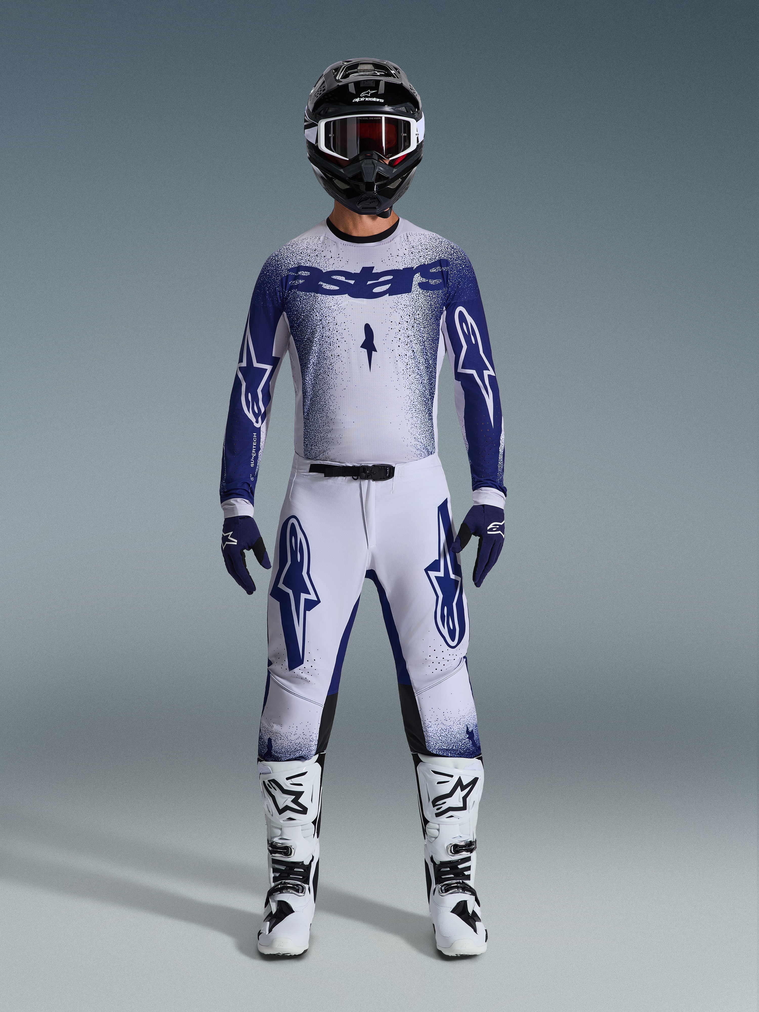 Motocross rider wearing the Alpinestars 2026 Supertech Scenz Gear Set, white and dark blue jersey with matching pants, features large Astars logos and speckled graphic design, paired with a black helmet, white and black protective boots, and coordinated blue gloves, complete professional racing outfit