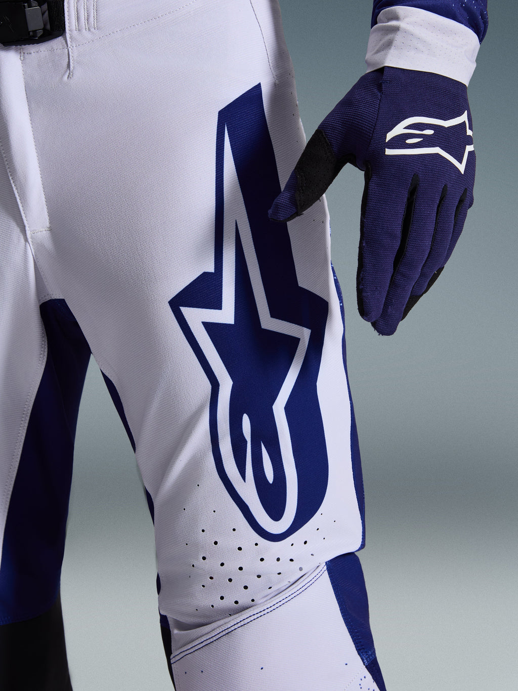 Alpinestars 2026 Supertech Scenz Pants, calças de motocross, cinzento claro e azul escuro, vista de pormenor da coxa com logótipo Alpinestars grande em azul escuro, zonas de ventilação perfuradas e tecido rip-stop elástico, usadas com uma luva de motocross azul escuro a combinar