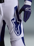 Alpinestars 2026 Supertech Scenz Pants, calças de motocross, cinzento claro e azul escuro, vista de pormenor da coxa com logótipo Alpinestars grande em azul escuro, zonas de ventilação perfuradas e tecido rip-stop elástico, usadas com uma luva de motocross azul escuro a combinar
