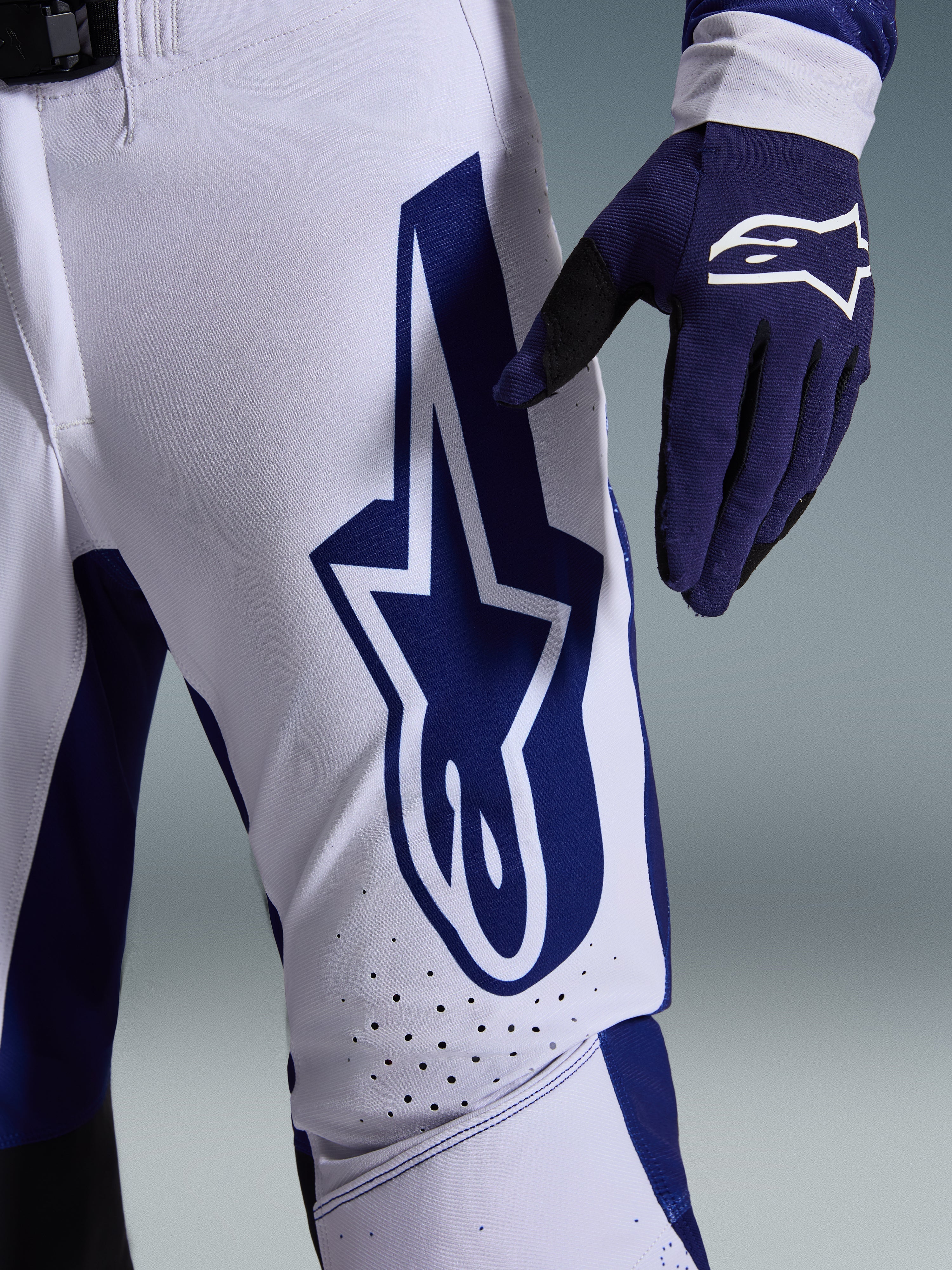 Alpinestars 2026 Supertech Scenz Pants, calças de motocross, cinzento claro e azul escuro, vista de pormenor da coxa com logótipo Alpinestars grande em azul escuro, zonas de ventilação perfuradas e tecido rip-stop elástico, usadas com uma luva de motocross azul escuro a combinar