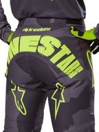 Alpinestars 2025 Racer Hollow Pants, calças de motocross, camuflagem escura e amarelo néon, com um grande logótipo Alpinestars amarelo na parte traseira, padrão de camuflagem tática com detalhes amarelos vibrantes e construção durável para corridas todo-o-terreno.