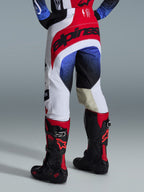 Alpinestars 2026 Techstar Knif Pants, calças de motocross, brancas, vermelhas e azuis, vista traseira mostrando assento vermelho com logótipo preto, padrões salpicados a azul e preto nas pernas, usadas com botas Tech 10 pretas e vermelhas, equipamento de competição todo-o-terreno de elite