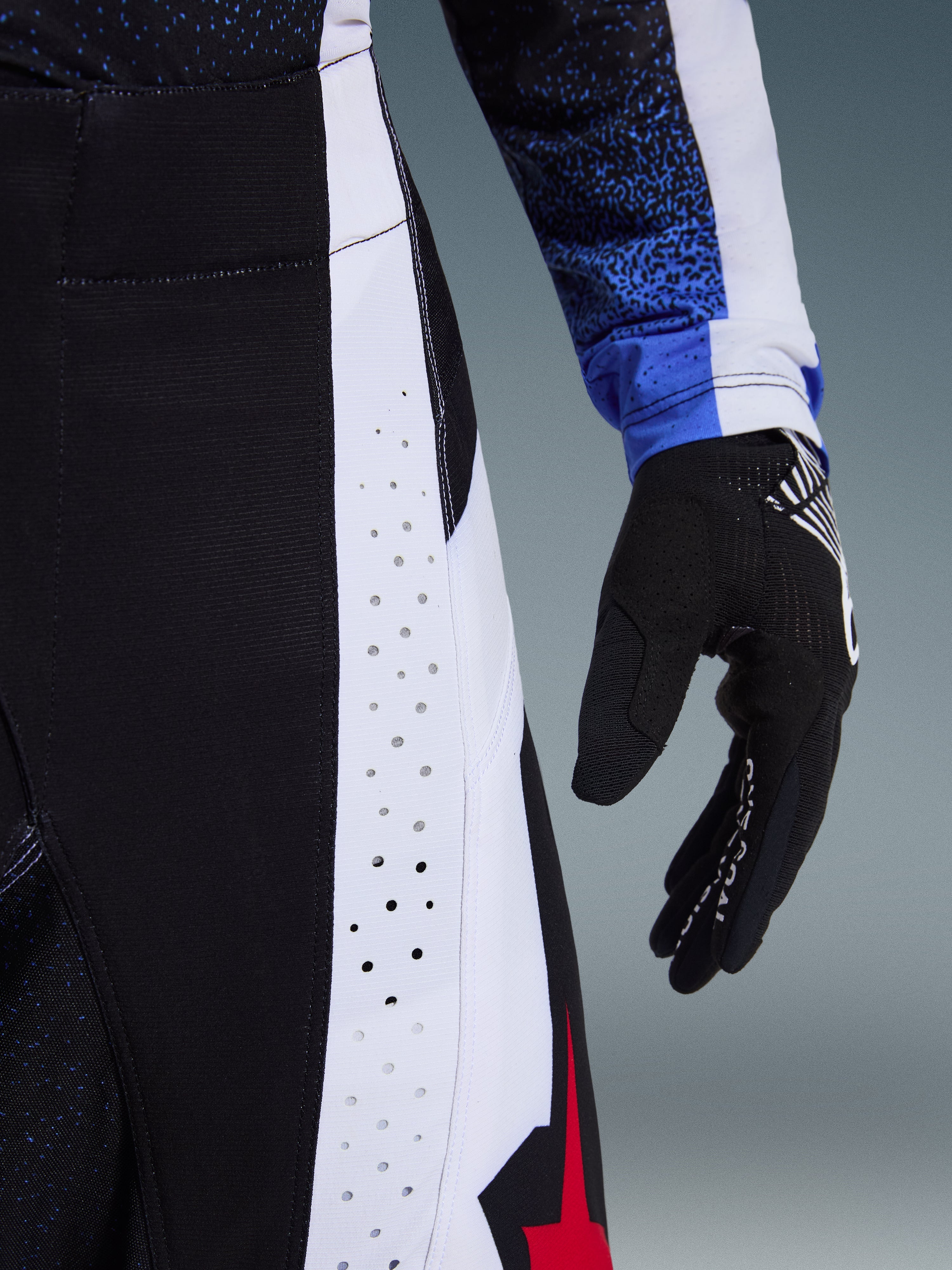 Pormenor das calças Alpinestars 2026 Techstar Knif em preto, branco e vermelho, destacando as perfurações cortadas a laser no painel lateral branco para ventilação, com luva de motocross preta e manga de camisola azul com padrão, evidenciando a construção de alta performance.