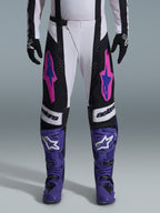 Piloto de motocross com o conjunto Alpinestars 2026 Techstar Knif, calças brancas e pretas com logótipos cor-de-rosa e azuis, botas roxas com logótipos brancos e luvas pretas, apresentando um corte atlético de competição e ventilação cortada a laser para corridas fora de estrada