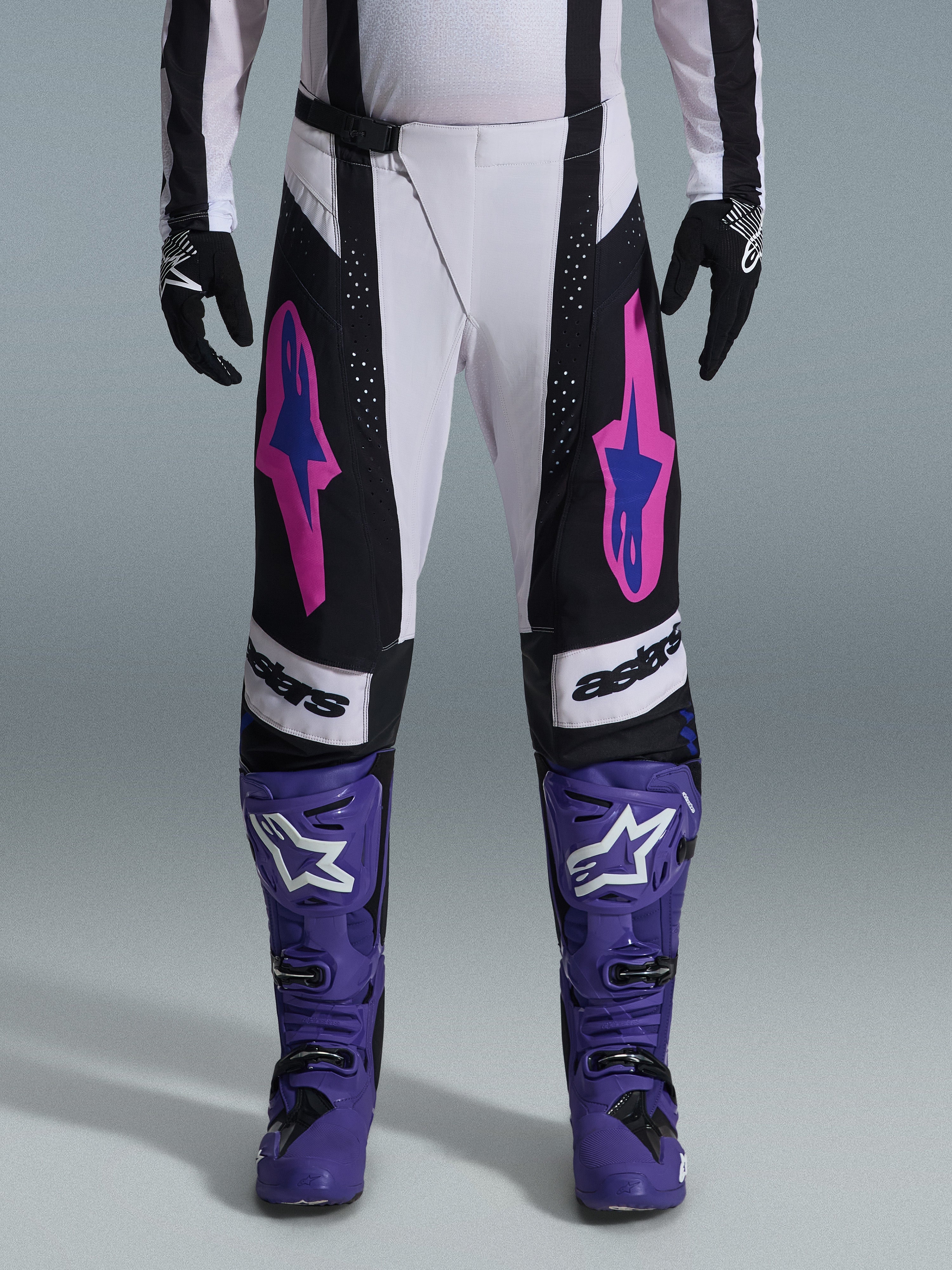 Alpinestars 2026 Techstar Knif Pants, calças de motocross, pretas, brancas e roxas com detalhes rosa e azul, usadas com botas de corrida roxas e luvas pretas, com corte de corrida ergonómico e grafismos de logótipo arrojados para desempenho em todo-o-terreno