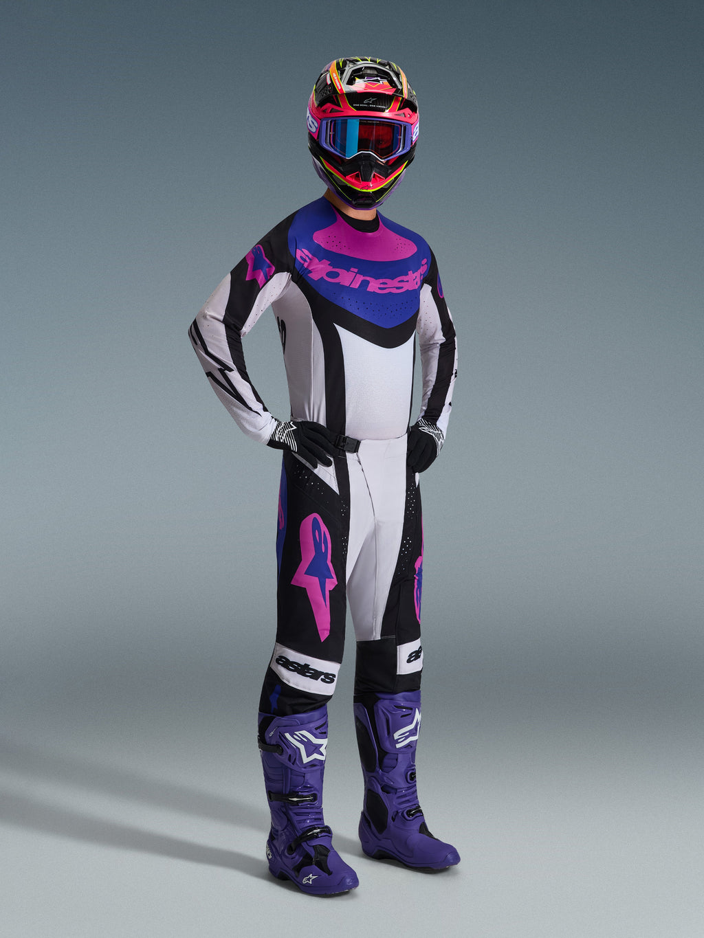 Piloto de motocross com o conjunto de equipamento Alpinestars 2026 Techstar Knif, camisola branca com detalhes em preto, azul e rosa, calças a condizer com logótipos Astars cor-de-rosa, capacete multicolorido com óculos, botas de corrida roxas, equipamento de alto desempenho coordenado para competição de motocross