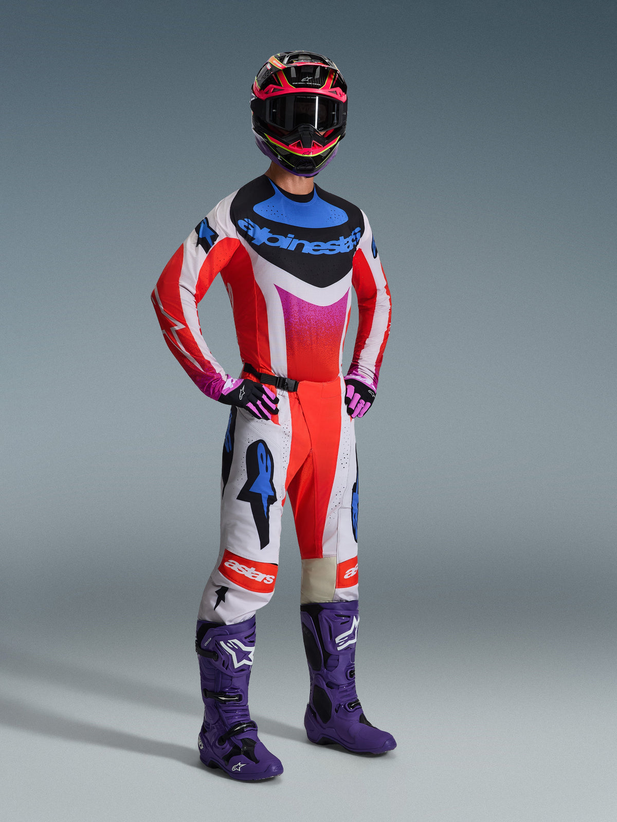 Alpinestars 2026 Techstar Knif Gear Set, piloto de motocross com equipamento de corrida completo, camisola vermelha com detalhes em branco e azul, calças brancas com logótipos de estrela vermelhos e azuis, botas de motocross roxas, capacete preto e cor-de-rosa, kit de corrida profissional para todo-o-terreno