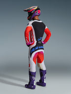 Motocross rider viewed from behind wearing complete racing gear, Alpinestars 2026 Techstar Knif Pants in Light Gray and Orange Fluo with blue branding, matching jersey with orange sleeves, purple Tech 10 boots, and a colorful racing helmet, coordinated professional off-road outfit